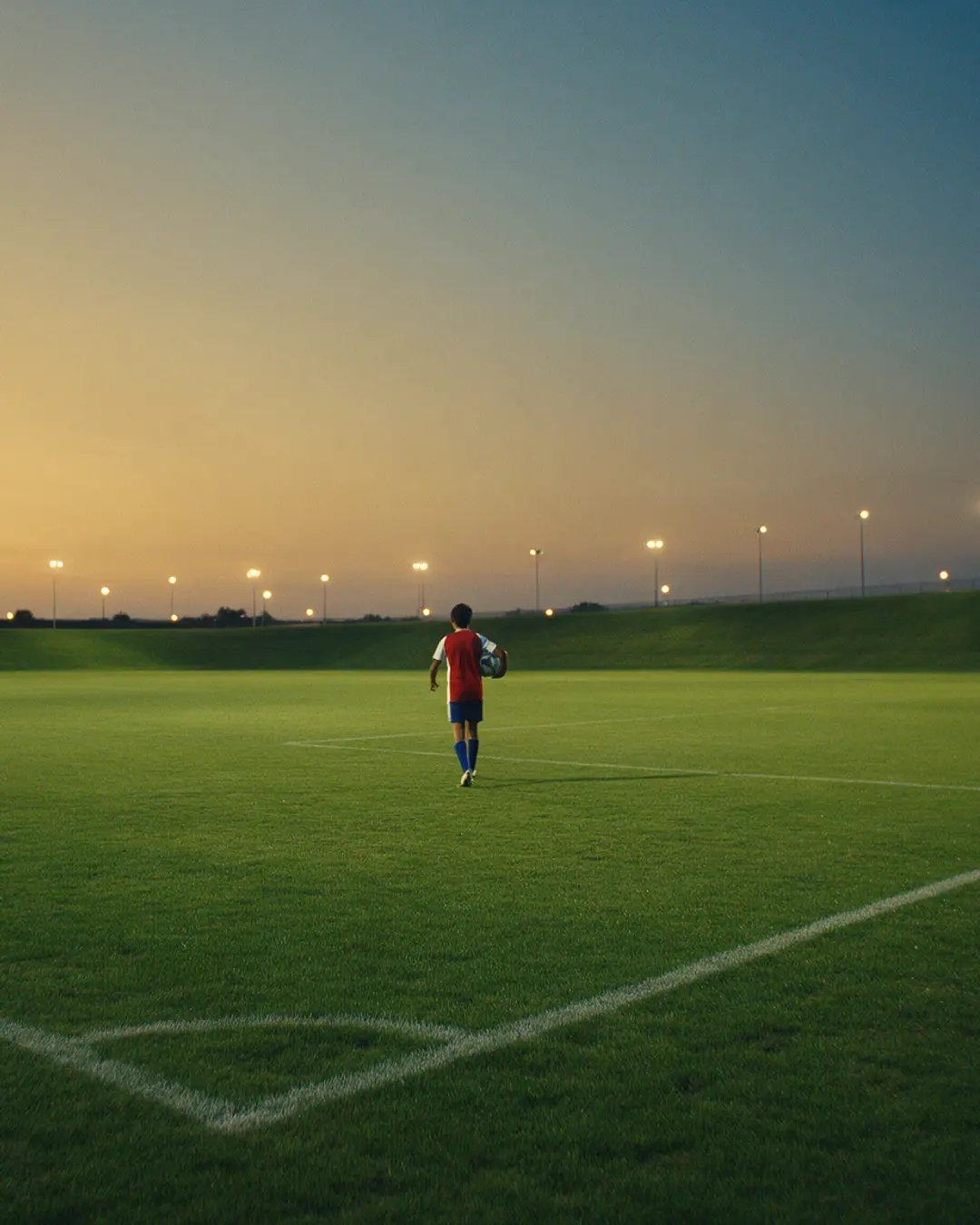Un joven futbolista camina con el balón por una cancha abierta al atardecer, con las luces del estadio de fondo.