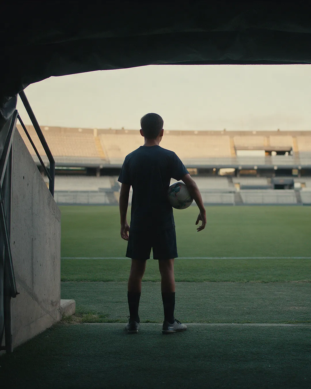 Un joven futbolista, de espaldas y con el balón bajo el brazo, mira una gran cancha desde la boca de un túnel al atardecer.