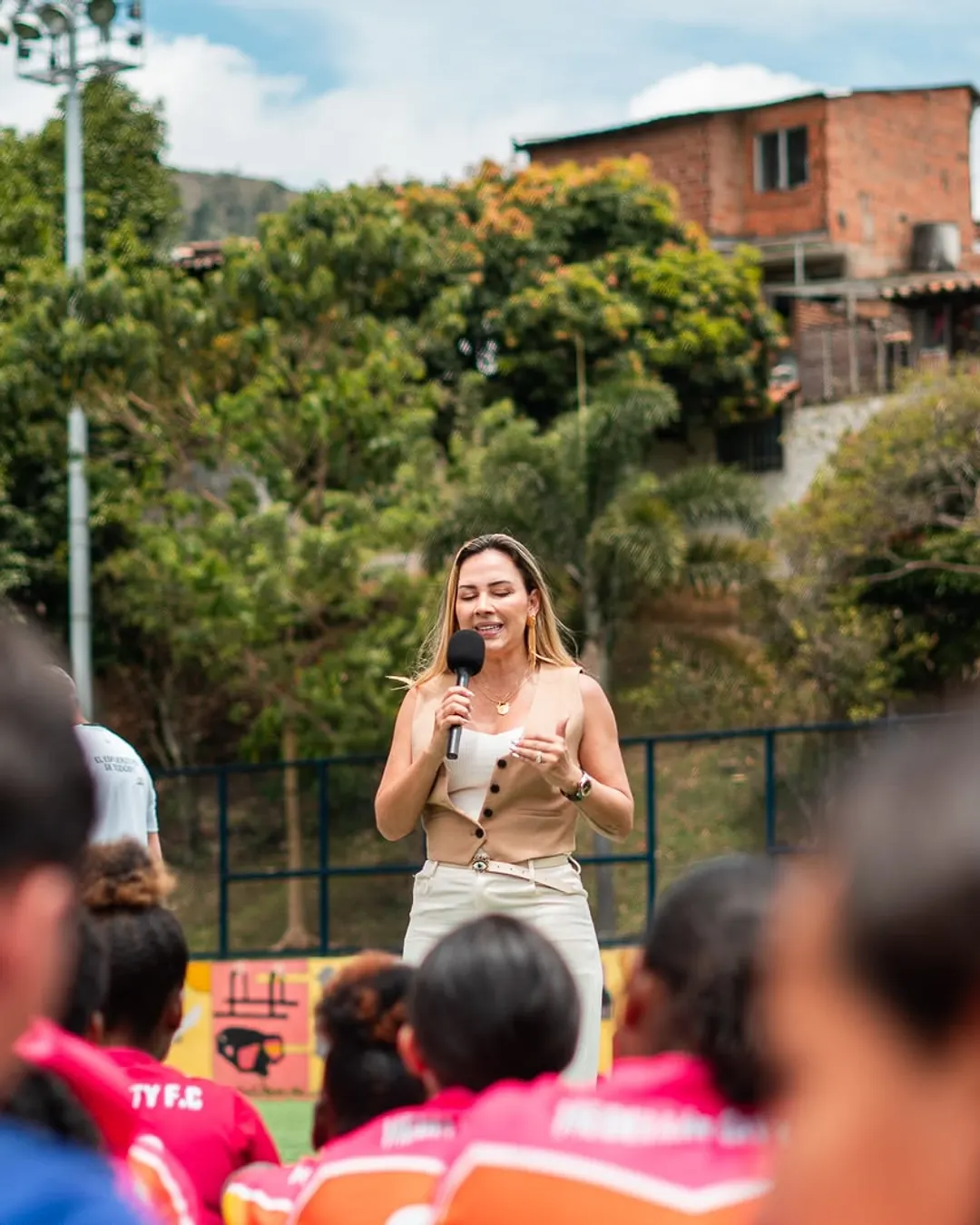 Una mentora de 11 Cracks habla con un micrófono frente a un grupo de jóvenes deportistas, en una cancha de barrio.