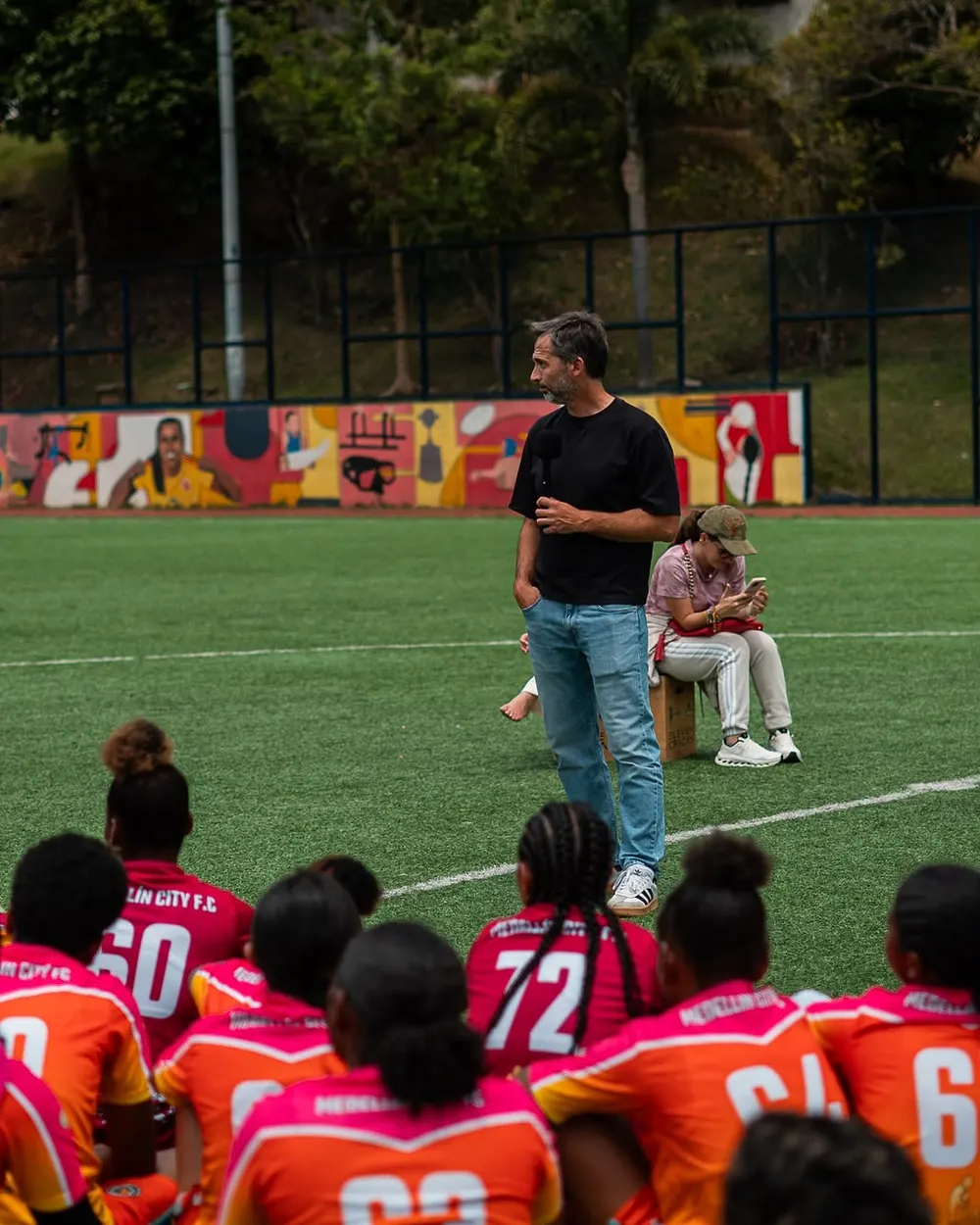 Un hombre habla con un micrófono frente a un equipo juvenil sentado en la cancha, en un encuentro real entre personas de distintos entornos deportivos.